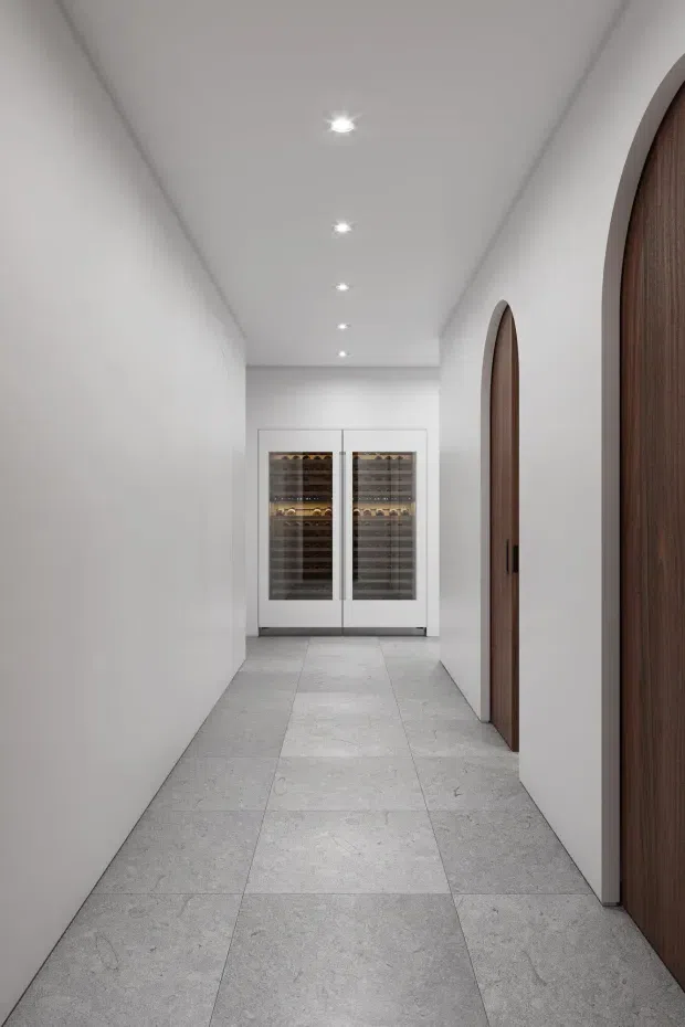 A narrow, minimalist interior hallway with white plaster walls and ceiling, recessed downlights, large pale grey stone-look floor tiles, and tall arched timber doors along the right side. At the end of the corridor are two built-in glass-fronted wine refrigerators or wine cellars, creating a refined residential wine-storage area.