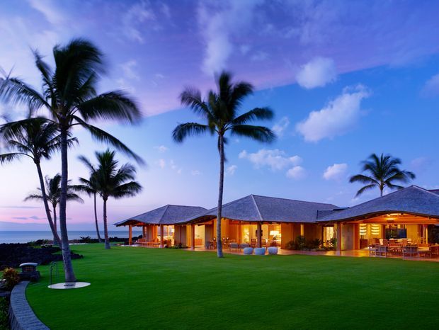 A low-slung tropical resort-style residence viewed from a broad manicured lawn at dusk. The single-storey building has multiple hipped roof forms with dark grey roofing, warm timber-toned columns and illuminated interiors, generous open-sided living areas, and extensive glazing. Tall palm trees frame the property, with a curved paved walkway across the lawn and a visible ocean horizon beyond.