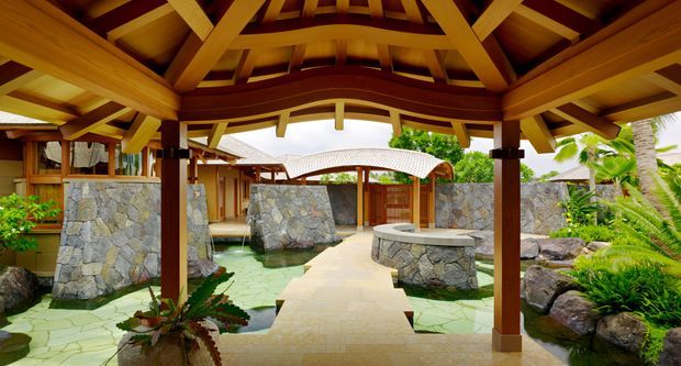 Open-air covered pavilion with a dramatic exposed timber roof structure, overlooking a landscaped tropical courtyard. A pale stone pathway crosses shallow water features toward a rounded stone seating or bar structure, with rugged stone walls, timber buildings, lush palms, ferns, and tropical planting throughout.