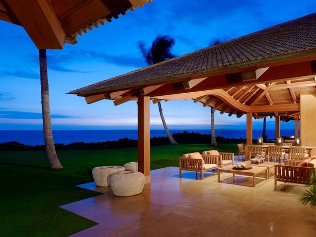 Open-air tropical resort-style pavilion or terrace with substantial timber posts and exposed timber rafters, a tiled pitched roof, polished stone or tile flooring, and warm wooden lounge and dining furniture. The space overlooks a dark ocean horizon and landscaped lawn, with palm trees and a vivid blue dusk sky beyond.