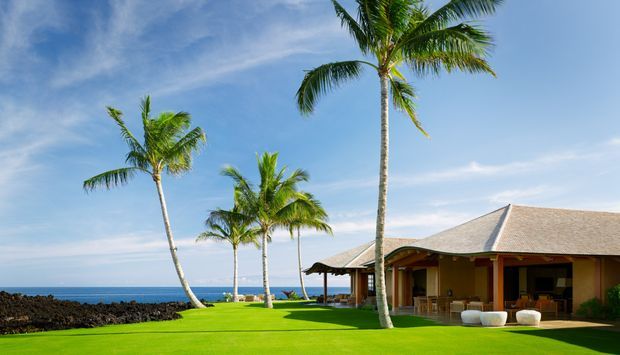 A tropical coastal resort-style pavilion beside the ocean, with a low hipped roof, open glazed living area, broad lawn, and tall palm trees. The image is brightly lit with a relaxed beachfront setting and clear blue sky.
