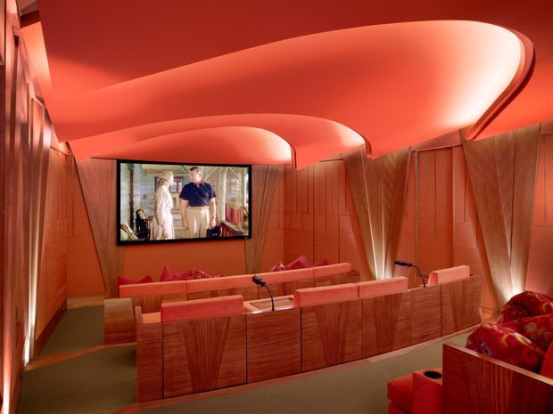 A richly detailed home cinema with tiered red upholstered seating, a wall-mounted screen, sculptural curved illuminated ceiling panels, and warm wood wall panelling and cabinetry. The space has a dramatic, immersive red-orange lighting scheme and integrated architectural detailing.