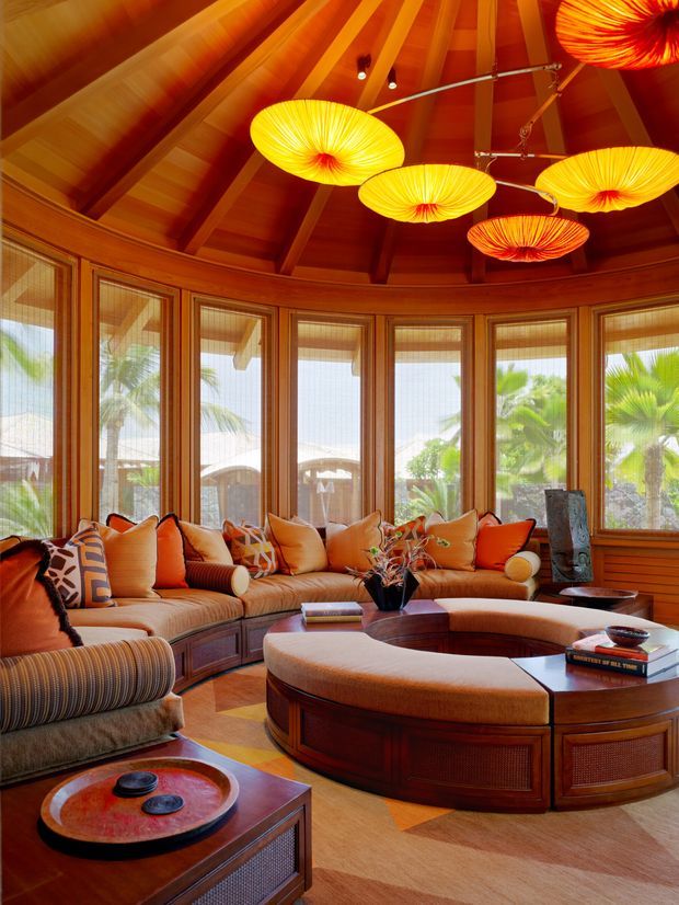 A richly furnished, curved sunroom or living room with extensive timber-framed windows, warm wood-panelled walls and a dramatic exposed radial timber ceiling. A large curved sectional sofa surrounds an unusual custom oval coffee table, while colourful sculptural pendant lights hang overhead. Tropical palms are visible outside through the glazing.