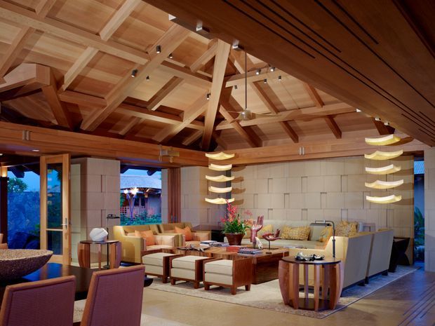 Spacious, open-plan living room with extensive warm timber detailing, exposed pitched roof trusses, clerestory-like glazing, and a large modular seating arrangement. The room includes lounge chairs, sofas, a central timber coffee table, sculptural pendant lights, a stone feature wall, and timber-framed openings to the exterior. The atmosphere is refined, warm, and resort-like.