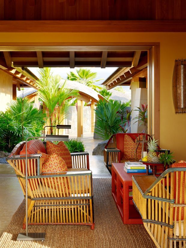 A richly coloured, open-sided tropical lounge or living area with timber beams and ceiling, surrounded by lush palms and connected to a landscaped courtyard. Woven rattan lounge chairs with patterned red-orange cushions are arranged around a red coffee table, creating a relaxed resort-like atmosphere. The space features warm timber surfaces, textured natural flooring, and strong indoor-outdoor connection.