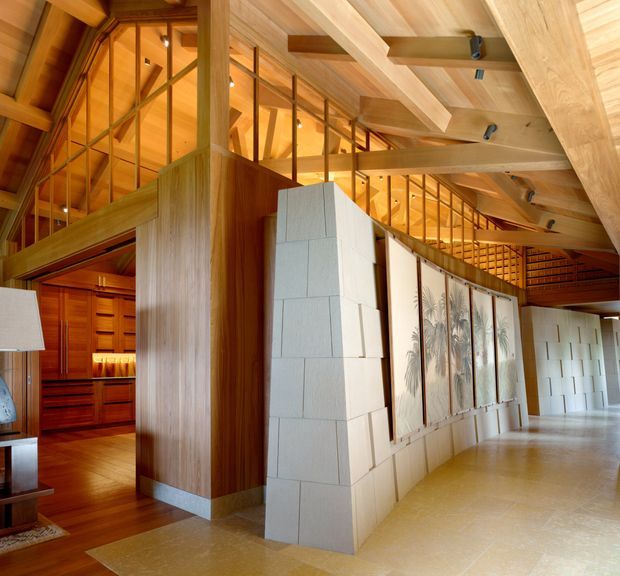 A spacious, warm-toned interior with extensive timber construction, exposed beams, clerestory-style glazing, and a mezzanine walkway. A sculptural pale masonry partition and framed artwork divide the open circulation area from a timber-lined room with built-in joinery. The space has a high, visually complex ceiling and strong natural wood character.