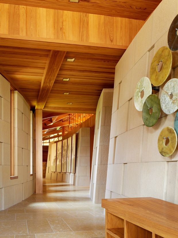 A long, warmly lit interior corridor with a richly finished timber ceiling, exposed structural beams, pale stone or rendered wall panels, and large stone floor tiles. Recessed ceiling lights lead toward a brighter timber-framed area at the far end. Decorative circular wall art and a built-in timber console add visual interest along the right wall.