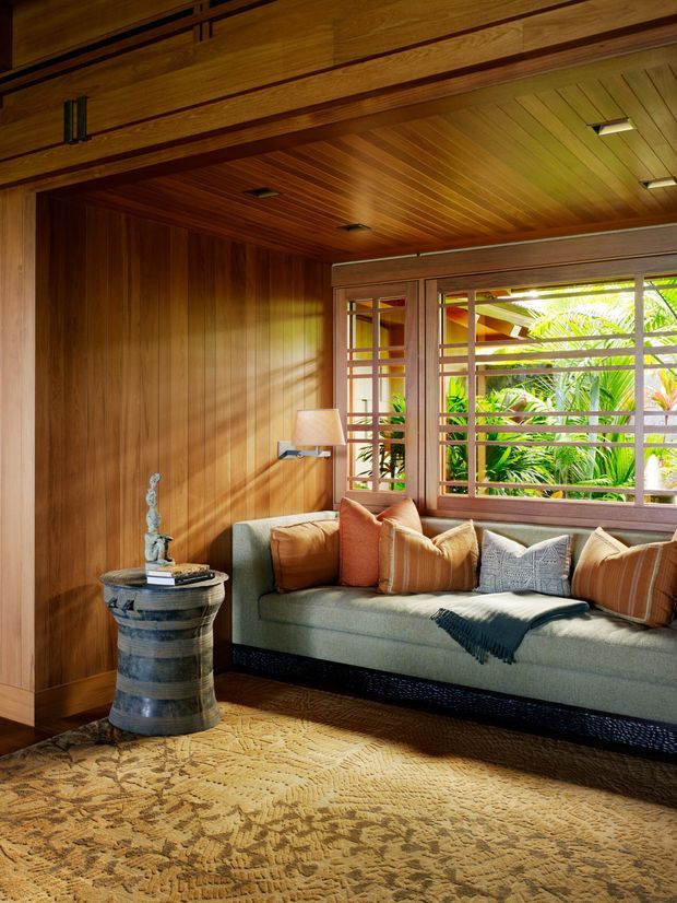 Warm, richly timber-lined living room with a built-in upholstered window seat beneath large multi-pane windows. The room includes a sculptural pedestal side table, table lamp, patterned rug, and layered neutral and rust-toned cushions. Tropical greenery is visible outside through the windows.