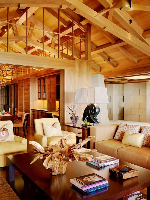 Warm, expansive living room with extensive timber construction, exposed beams, a pitched timber ceiling, and a glazed upper-level partition or mezzanine. The room contains cream upholstered seating, a large dark timber coffee table, decorative driftwood, built-in timber cabinetry, and a sculptural lamp. A dining area and bar cabinetry are visible beyond the living space.