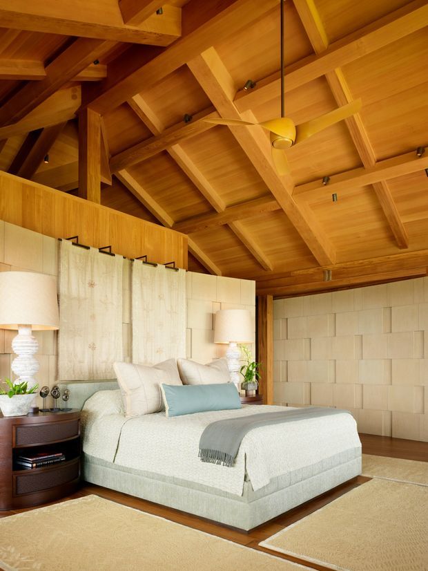 Warm, refined bedroom with a large upholstered bed, pale bedding, bedside tables and lamps, layered curtains, and a prominent exposed timber vaulted ceiling with structural beams and a ceiling fan. The walls feature light block-patterned paneling, creating a calm natural retreat.