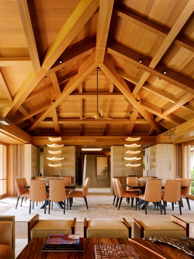 A richly timber-lined open-plan dining and living space beneath a dramatic exposed pitched roof. Large timber trusses and rafters dominate the ceiling, with two dining tables surrounded by woven tan dining chairs, low lounge seating in the foreground, pale masonry walls, and sculptural suspended pendant lights. The room has a warm, resort-like, tropical architectural character with abundant natural timber and daylight.