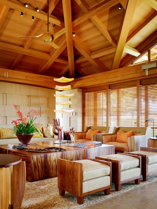 Warm, richly timber-lined living room with a dramatic exposed post-and-beam vaulted ceiling, integrated spot lighting, ceiling fan, and extensive horizontal timber blinds. The room contains a large central wood coffee table, upholstered lounge chairs and sofas in orange and cream tones, a patterned area rug, decorative objects, and a sculptural hanging light. High-level glazing and clerestory windows bring in natural light.