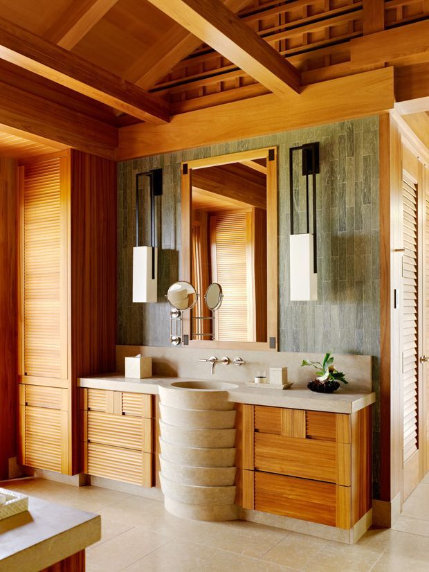 Warm, richly detailed bathroom featuring a sculptural stacked-stone pedestal basin, pale stone vanity countertop, timber cabinetry, louvred timber doors, and a large framed mirror. The walls are clad in vertically textured green-grey stone tiles, while exposed timber beams and a slatted timber ceiling create a strong natural, resort-like character. Twin wall sconces and polished bathroom fittings flank the mirror.
