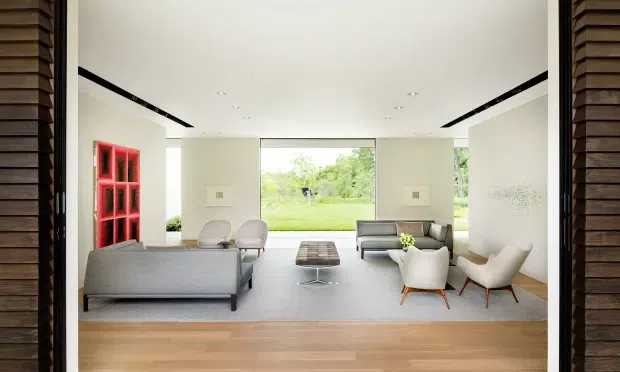 Bright contemporary living room viewed through a wide opening, with a grey sofa, two light lounge chairs, a dark coffee table, pale carpeting, and timber flooring in the foreground. White walls and ceiling with recessed downlights frame large full-height glazing overlooking a green landscape. A vivid red built-in shelving or display feature adds contrast, while dark horizontal timber cladding is visible around the opening.