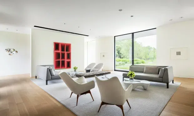 Bright, spacious contemporary living room with pale walls, broad timber flooring, a large light rug, sculptural cream lounge chairs, grey sofas, a low organic-shaped coffee table, and a striking red built-in display shelf. Floor-to-ceiling glazing brings views of greenery into the room, while recessed ceiling lighting and minimal detailing create a refined, airy interior.