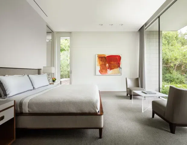 Bright, spacious contemporary bedroom with a low-profile upholstered bed, crisp white bedding, and neutral carpeting. Floor-to-ceiling glazing and a full-height mirrored sliding partition open the room to leafy views, while a pair of upholstered lounge chairs and a small glass table create a sitting area. Minimal white walls, recessed ceiling lights, and a colorful abstract artwork complete the calm, refined interior.