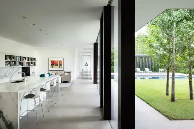Bright, open-plan contemporary kitchen and living space with a long white marble island, integrated sink, and white bar stools. Floor-to-ceiling black-framed glazing and strong vertical structural elements connect the interior to a landscaped lawn and swimming pool outside. Built-in shelving, recessed ceiling lights, pale flooring, and minimal furnishings create a clean, airy interior.
