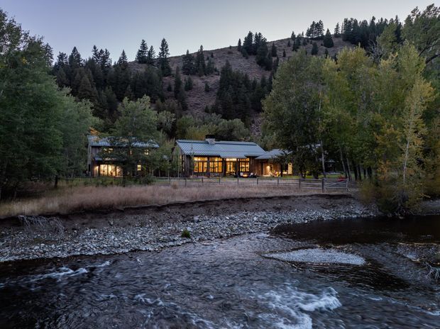 A single-storey house nestled in a forested mountain setting beside a rocky flowing river. The home has a low-pitched metal roof, extensive warm-lit glazing, dark exterior finishes, and a long, low horizontal form integrated into the wooded landscape.