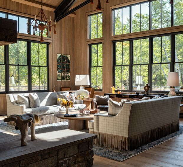 A spacious, warmly lit living room with extensive floor-to-ceiling black-framed glazing overlooking trees. The room features vertical timber wall and ceiling panelling, a large stone fireplace hearth in the foreground, tailored upholstered sofas and armchairs, a round timber coffee table, layered rugs, table lamps, and a sculptural hanging light. The overall look is refined rustic with strong natural materials and an indoor-outdoor connection.