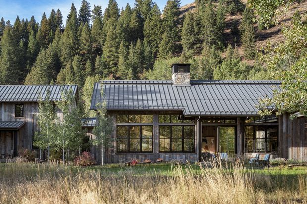 Exterior view of a low, contemporary mountain home set against a dense conifer forest. The building features dark vertical timber cladding, extensive black-framed glazing, and a standing-seam metal gabled roof with a stone-clad chimney. Native-style landscaping and dry grasses surround the house.