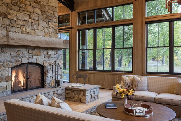 A warm, rustic living room centered around a substantial natural-stone fireplace with a lit fire. The room features extensive dark timber-framed glazing, timber wall panelling, a stone hearth and low table, upholstered sofas, and views into a wooded landscape.