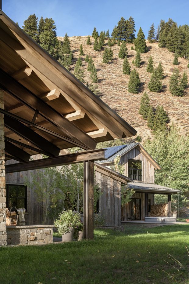 Exterior view of a rustic contemporary mountain home set against a wooded, dry hillside. The composition highlights broad timber roof overhangs with exposed rafters, dark structural beams, weathered vertical timber cladding, stone walls, large black-framed glazed openings, and a connected gabled wing.