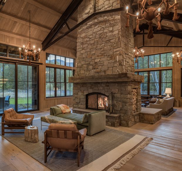 A spacious rustic living room centred around a large rough-stone masonry fireplace with a lit fire. The room features exposed timber ceilings and beams, expansive black-framed windows and glass doors, leather lounge chairs, sofas, area rugs, and warm chandelier lighting. Forested landscape is visible through the glazing.