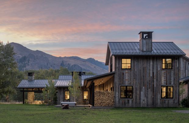 Exterior view of a rustic contemporary mountain residence at dusk, with weathered vertical timber cladding, dark standing-seam metal gable roofs, black-framed windows, covered outdoor connections, and neatly stacked firewood. A smaller matching outbuilding sits to the left, with wooded mountains forming the background.