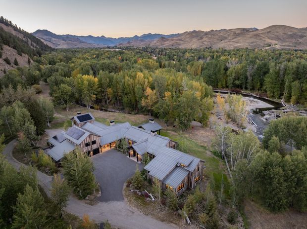 Aerial view of a sprawling multi-wing residence nestled in dense autumn woodland beside a winding river, with rugged mountain ranges in the background. The home has grey metal pitched roofs, extensive glazing, timber-toned exterior walls, and a dark paved driveway, forming a low-rise lodge-like complex integrated into the natural landscape.
