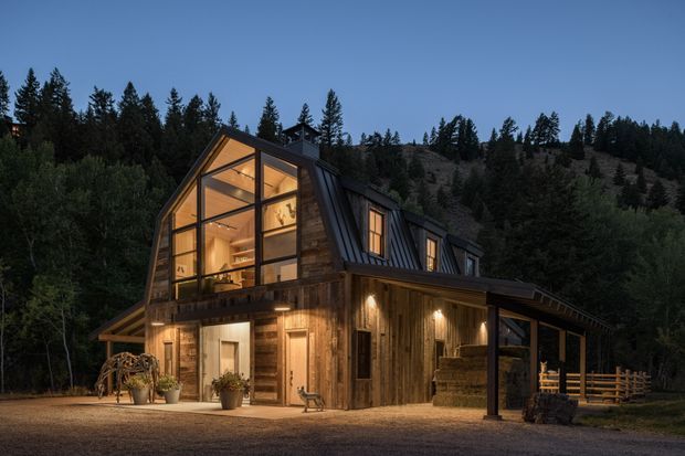 A warmly lit two-storey timber-clad mountain cabin surrounded by dense conifer forest. The steep gabled form features expansive upper-level glazing, dark metal roof sections, exposed timber framing, a covered outdoor porch, and illuminated entry doors.