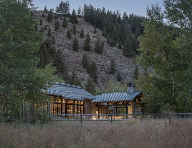A contemporary mountain lodge nestled in a wooded alpine landscape, with extensive floor-to-ceiling glazing, a dark metal pitched roof, timber structure and warm interior lighting. The building sits amid mature trees and dry meadow vegetation, backed by a steep forested hillside.
