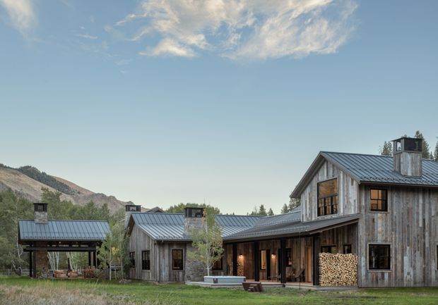 A large rustic timber-clad mountain residence with multiple connected pitched-roof volumes, dark metal roofing, timber-framed windows, covered outdoor areas, and stacked firewood beside the building. The home sits in a rural, mountainous landscape beneath a clear sky.