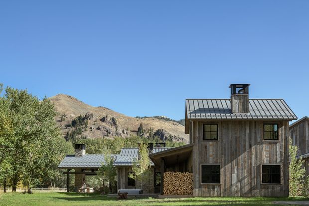 Exterior view of a rustic mountain property with weathered vertical timber cladding, dark metal pitched roofs, chimney, covered outdoor area, and stacked firewood. Mature trees, lawn, and rugged mountains form the surrounding landscape under a clear blue sky.