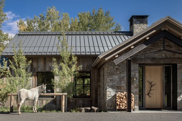 Exterior view of a rustic mountain-style home with a grey standing-seam metal roof, natural stone walls, timber-framed glazing, and a gabled entrance porch. A wood front door with a branch motif and stacked firewood reinforce the lodge-like character, while surrounding mature trees and a horse suggest a rural or alpine setting.