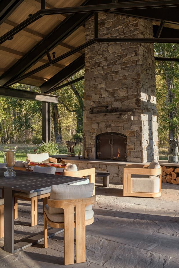 Open-air covered outdoor living and dining area set within a wooded landscape. The space features a large rustic stone fireplace with timber mantel and firewood storage, exposed dark steel framing, a warm timber-lined pitched ceiling, expansive glazing, and a slate or flagstone floor. A substantial timber dining table and upholstered chairs occupy the foreground, with lounge seating arranged around the fireplace.