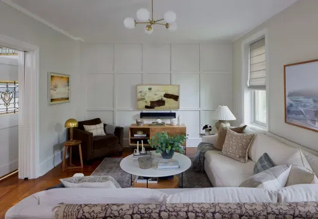 Bright, traditionally styled living room with white paneled walls, warm timber flooring, a large cream sectional sofa, brown lounge chairs, a round coffee table, layered rugs, framed artwork, and a brass globe chandelier. Tall windows bring in natural light, while a stained-glass doorway is visible at the left.