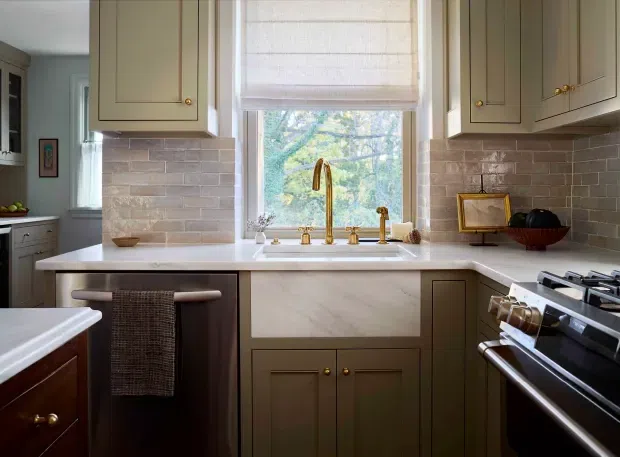 A warm, traditional kitchen with muted taupe-grey Shaker cabinetry, brass knobs and plumbing fixtures, a light stone benchtop, and a glossy pale brick-pattern tiled splashback. The compact layout includes a peninsula with seating, a farmhouse-style sink beneath a window with a woven Roman blind, and a stainless-steel range on the right. Natural greenery is visible outside the window.