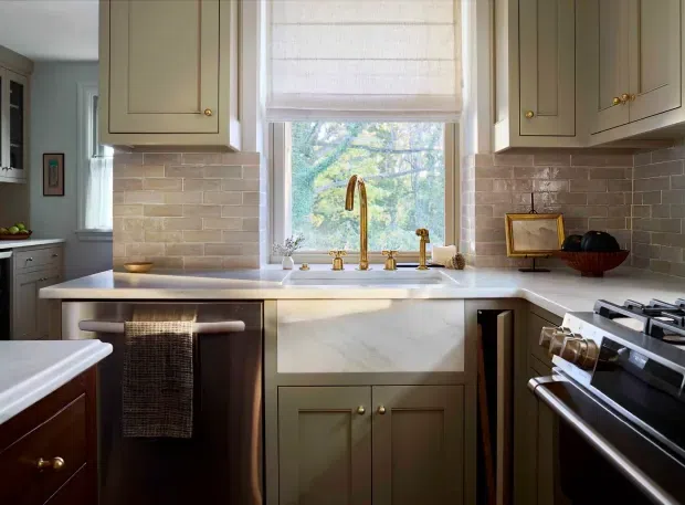 A compact, traditional-style kitchen with olive-grey shaker cabinetry, brass hardware, a light stone benchtop, and a pale brick-pattern tile splashback. The sink is positioned beneath a large window with a woven Roman blind, while a stainless steel range cooker occupies the right side. Warm natural light and timber accents create an inviting, refined atmosphere.