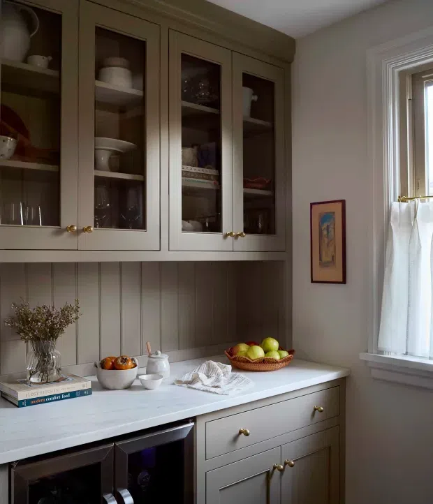 A softly lit traditional kitchen or butler’s pantry vignette with built-in taupe-grey cabinetry, glass-front upper cupboards displaying crockery and glassware, vertical panelled wall backing, and a pale countertop. Brass knobs, a fruit bowl, crockery, books, and a curtained window create a warm, refined domestic setting.