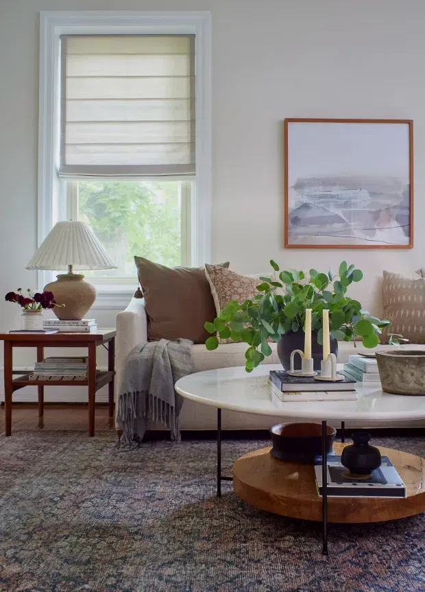 A warmly styled living room with a cream upholstered sofa, layered neutral cushions, a textured throw, round nesting coffee tables, a large patterned area rug, and a side table with a ceramic lamp. A large window has a light Roman shade, while framed abstract artwork and leafy indoor plants add softness and colour. The room has a refined, comfortable transitional feel with natural textures and muted earthy tones.