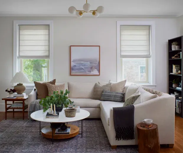 A bright, refined living room with a large L-shaped cream sectional sofa, layered neutral cushions, a round white coffee table, patterned area rug, side tables, indoor plants, and abstract artwork. Two tall windows with roman blinds provide natural light, while a sculptural globe chandelier and dark shelving add character. The room has a warm, contemporary-traditional feel.