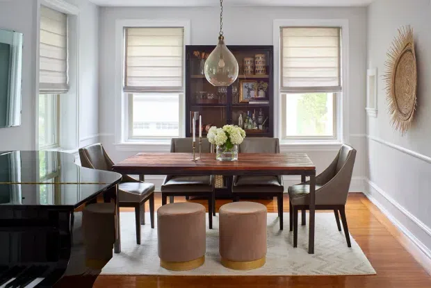 A refined dining room with a rectangular dark timber dining table, upholstered dining chairs, and two round ottomans arranged on a large rug. Pale grey walls with white trim and wainscoting surround three tall windows with roller blinds. A dark built-in or freestanding bar cabinet occupies the rear wall, while a sculptural pendant light hangs above the table. Warm timber flooring and restrained decorative accents create an elegant, inviting atmosphere.