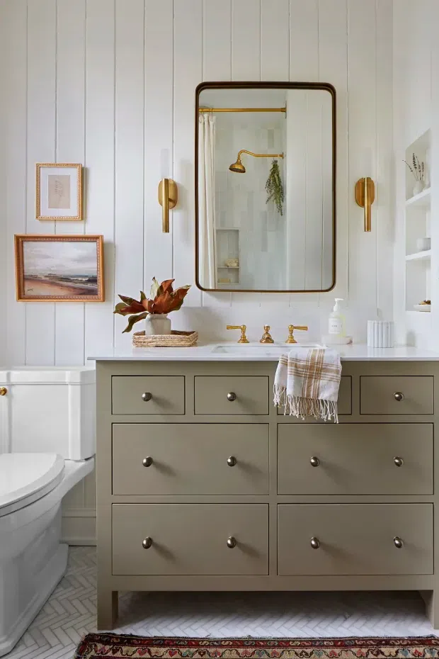 A thoughtfully styled bathroom featuring a large sage-grey double vanity with brass hardware and twin wall-mounted faucets, a white countertop, vertical white wall panelling, and a large rounded-rectangle framed mirror. A toilet is visible at left, while brass sconces, framed artwork, open shelving, decorative greenery, and a patterned runner add warmth and character. The floor is finished with small light herringbone tiles.