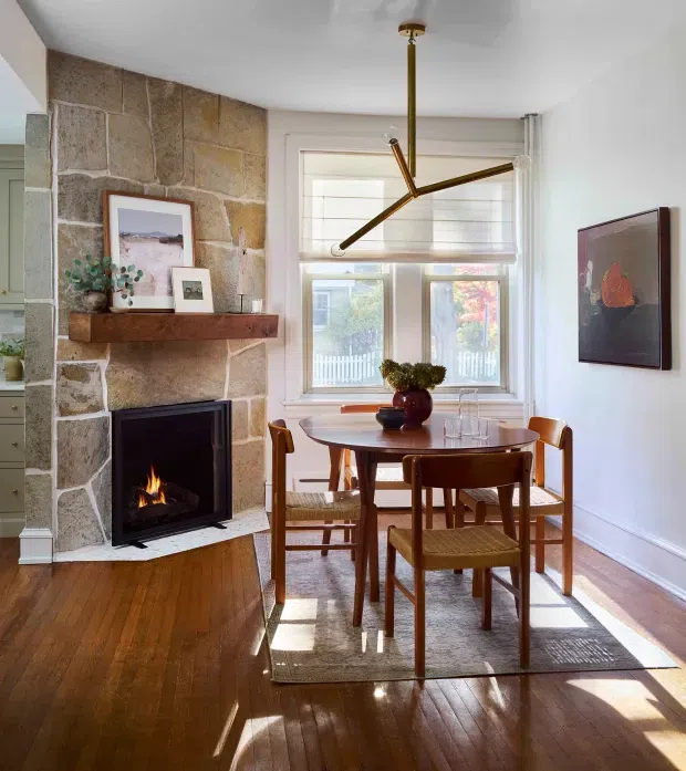 A bright, warmly styled dining area with a round wooden dining table and woven-seat chairs beside a substantial natural stone fireplace. The room features polished timber flooring, white walls and ceiling, large timber-framed windows with blinds, a sculptural brass-toned pendant light, and restrained artwork and decor. Sunlight creates strong highlights across the floor and rug.