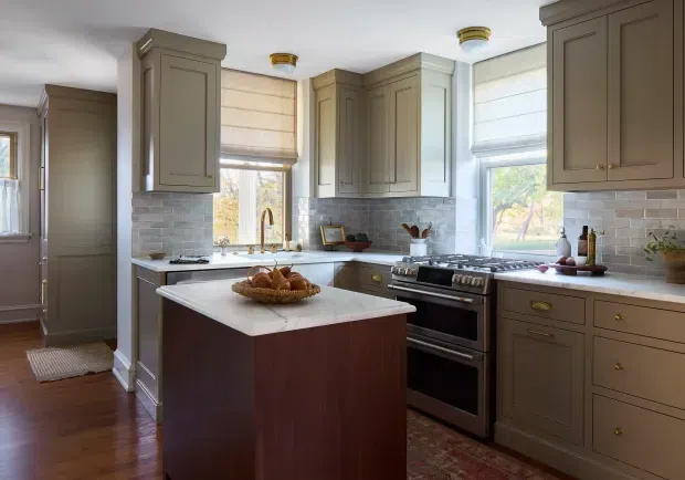 A traditional-style kitchen with taupe-gray raised-panel cabinetry, white stone benchtops, and a central island with a dark timber base. Pale subway-tile splashback, brass cabinet hardware and tapware, warm timber flooring, and large windows with Roman blinds create a bright, refined, classic interior. A freestanding stainless-steel range cooker sits beneath the cabinetry, with a small adjoining hallway or utility area visible at left.