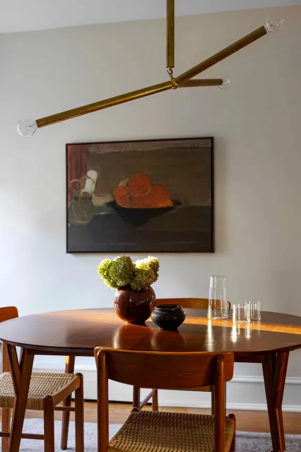 A warm, refined dining room with a polished mid-century wooden oval dining table and matching wooden chairs with woven cane seats. A sculptural brass linear pendant with exposed globe bulbs hangs above the table. The room has pale walls, timber flooring, a framed still-life artwork, and a simple table arrangement of flowers, ceramics, and glassware.