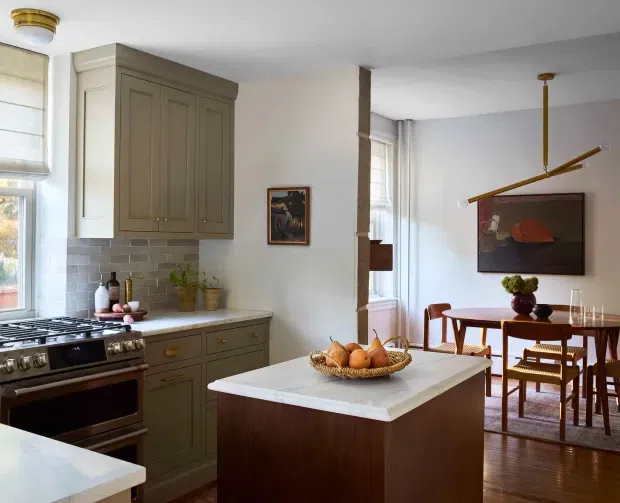 A warm, traditionally detailed kitchen opening into a dining area. The kitchen features muted grey-green shaker cabinetry with brass hardware, a pale tiled backsplash, a freestanding stainless-steel range, and a light stone island benchtop with a dark timber base. The adjacent dining area has a round timber table and chairs, timber flooring, framed artwork, and a sculptural brass-toned pendant light. Natural light enters through tall windows with Roman blinds.