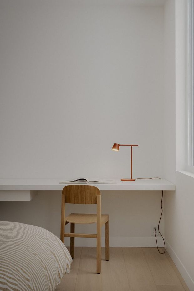 A minimalist bedroom corner with a built-in white wall-mounted desk, a simple light timber chair, an open book, and a slim copper desk lamp. The edge of a neatly made bed is visible in the foreground, while a white-framed window provides natural light. The room has pale walls and light timber flooring with a calm, uncluttered composition.