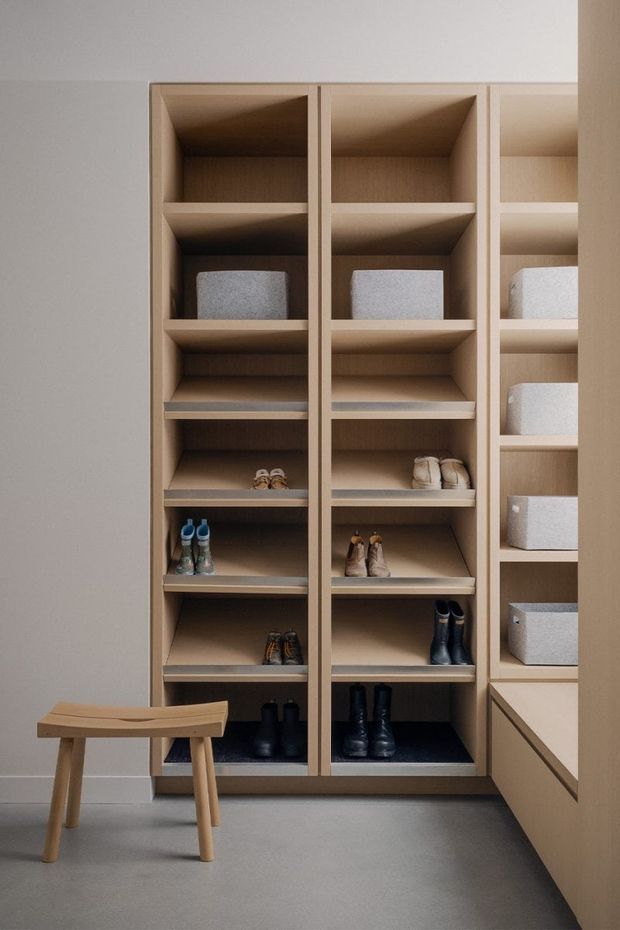 A minimalist built-in wardrobe and shoe-storage wall in a calm dressing or entry space. Full-height open timber shelving contains shoes, boots, and pale fabric storage boxes, with a small simple wooden stool in front. The cabinetry is framed by light neutral walls and sits above a smooth grey floor.