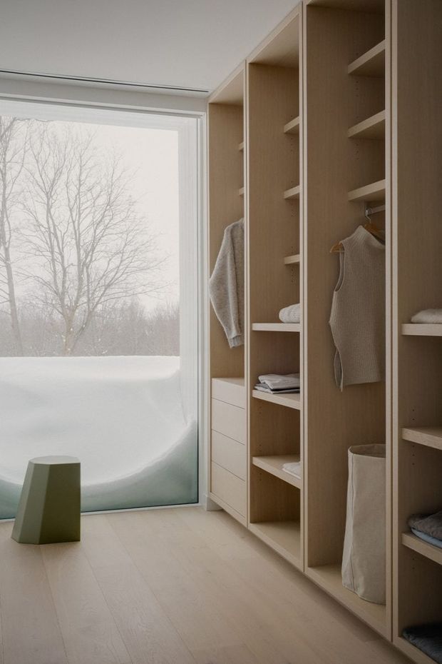 A minimalist walk-in wardrobe with full-height open timber shelving, hanging compartments, drawers, and neatly folded clothing. Pale timber flooring and warm light-wood joinery contrast with a large floor-to-ceiling glazed opening overlooking a snowy landscape. A small muted green stool sits near the glazing, creating a calm, restrained interior.